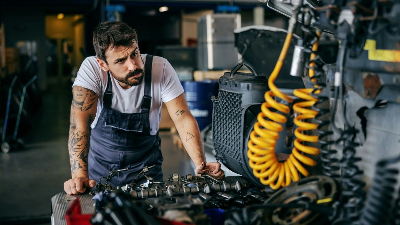 A man with tattoos and blue overalls leans over a table filled with mechanical parts.