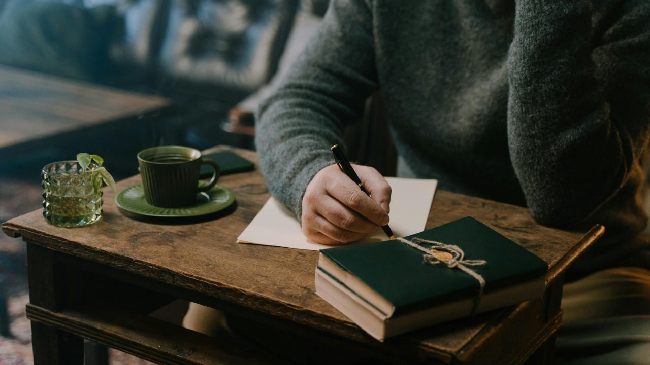 A person's hand writes with a pen on paper next to a green cup and book.