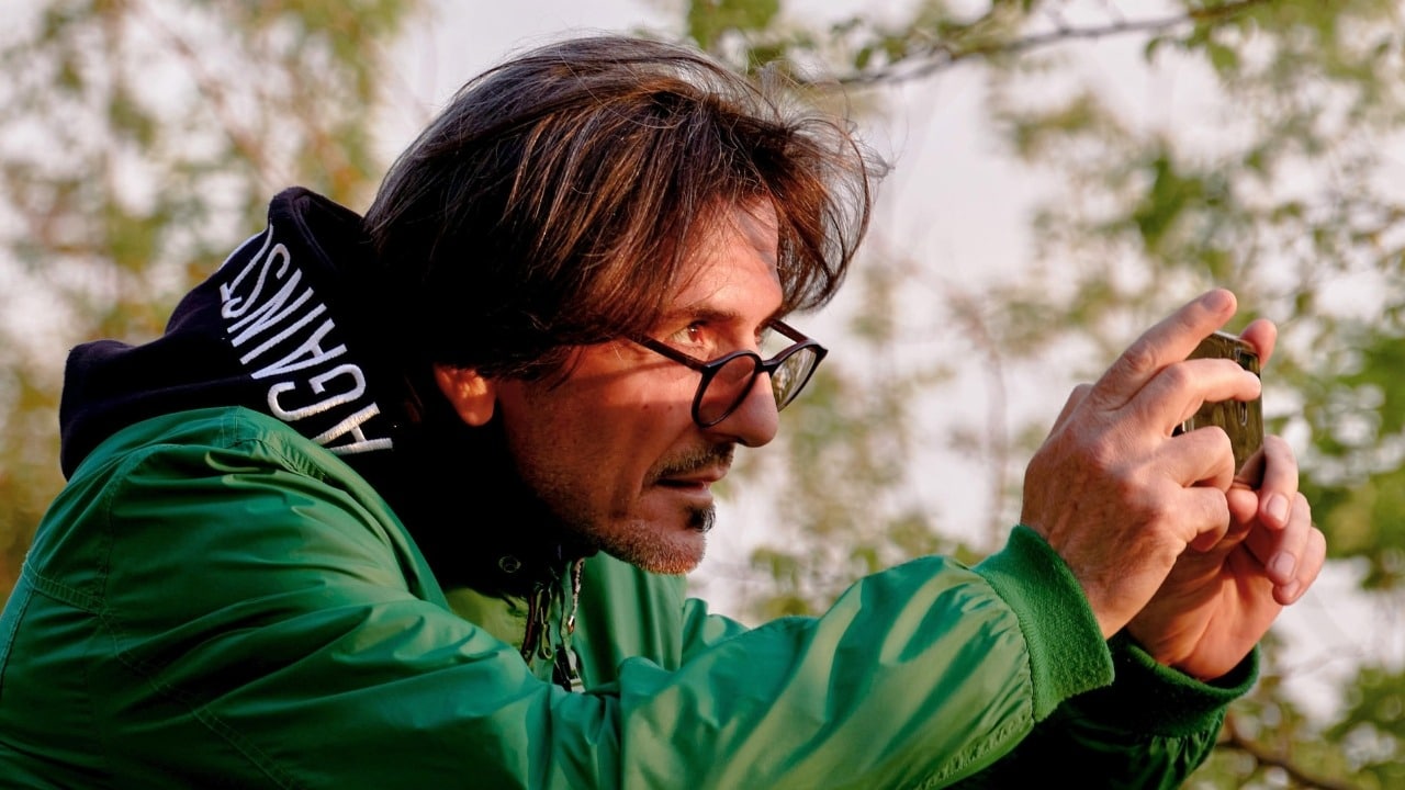 A man wearing a green jacket and glasses holds a smartphone up to take a photo.