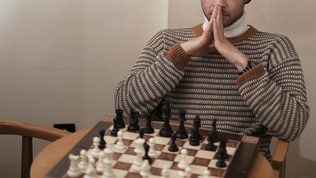 A man in a patterned sweater sits with his hands together behind a set-up chessboard.