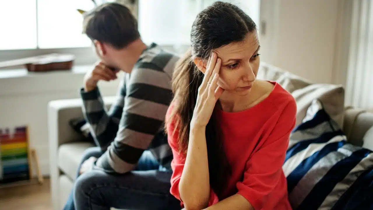 A woman in a red shirt sits on a sofa, touching her forehead with a frown.