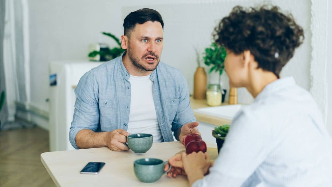 A man speaks with a tense expression while sitting at a table with another person.