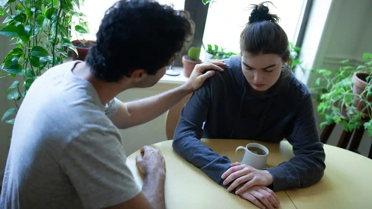 A person rests their hand on the shoulder of another person looking down at a table.