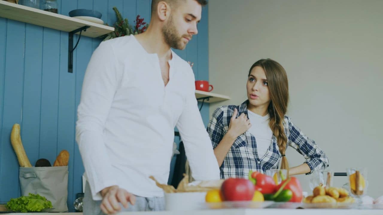 A woman in a plaid shirt speaks to a man while standing in a kitchen setting.