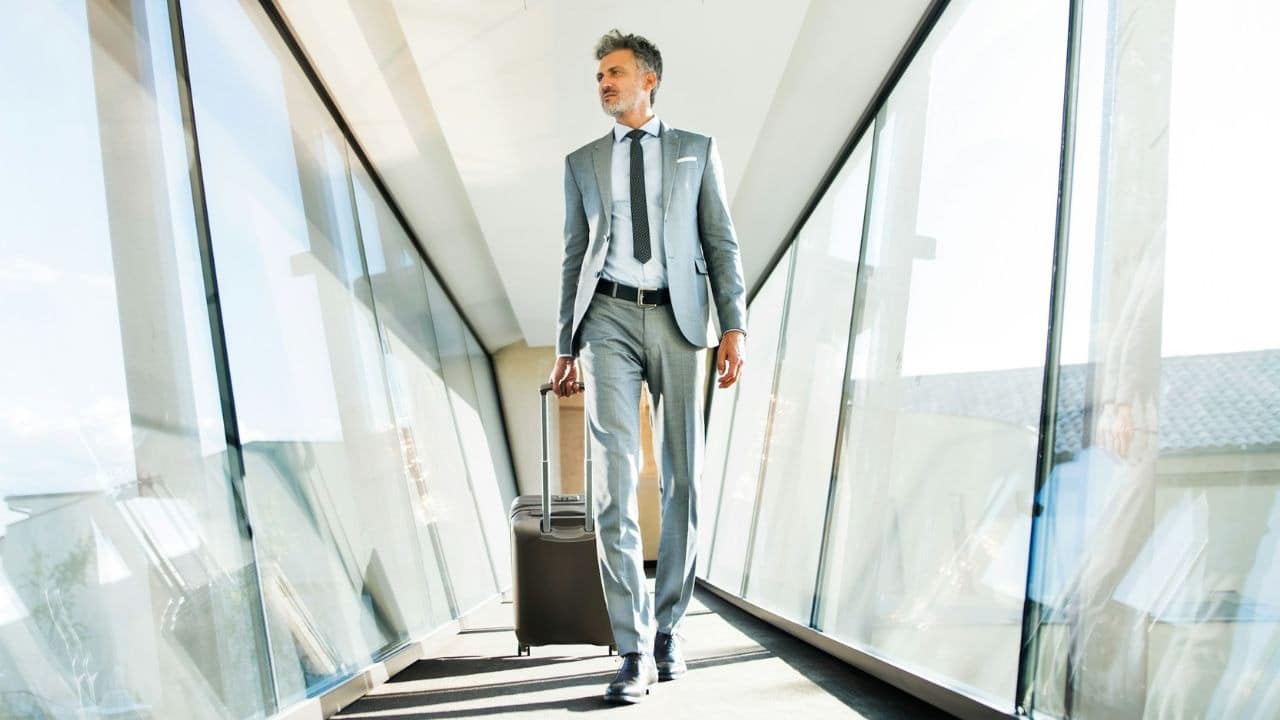 A man in a gray suit pulls a suitcase through a bright, glass-walled airport walkway.