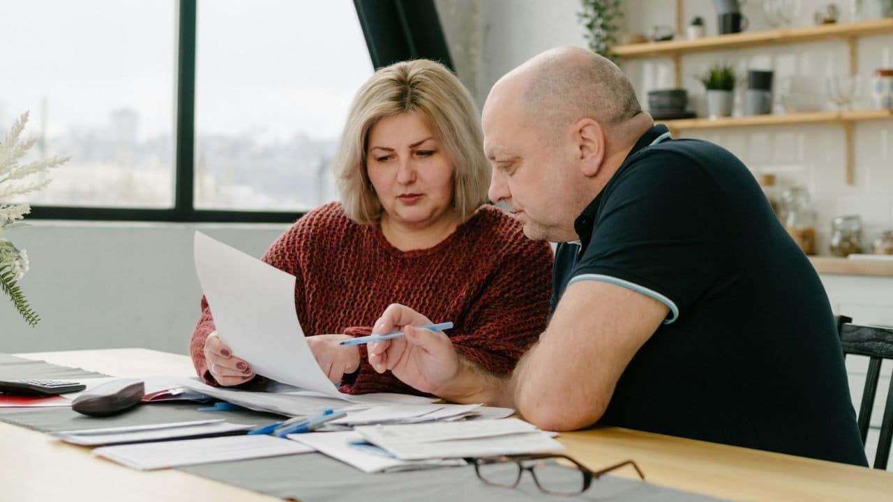 Two people sit at a wooden table together while reviewing several printed documents and papers.