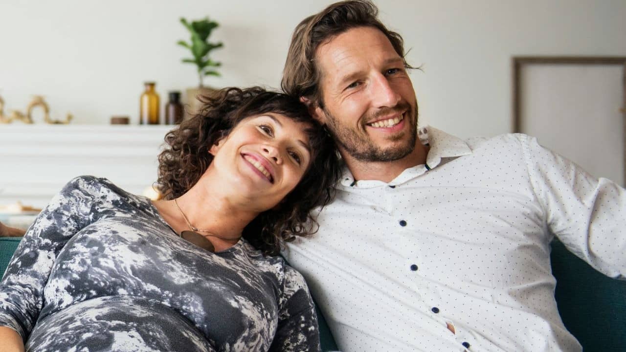 A smiling woman leans her head on a man’s shoulder while they sit on a couch.