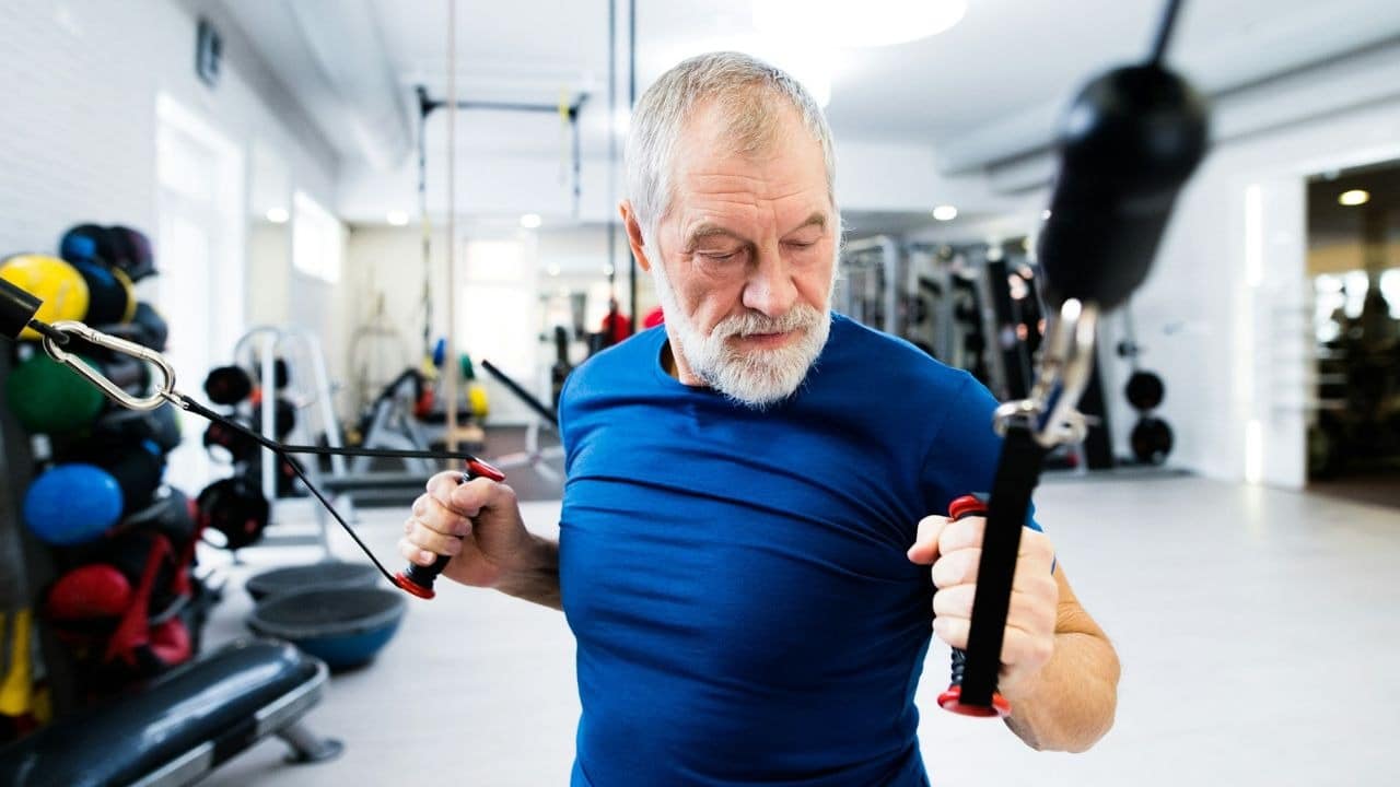 An older man with a white beard uses a cable machine while exercising in a gym.