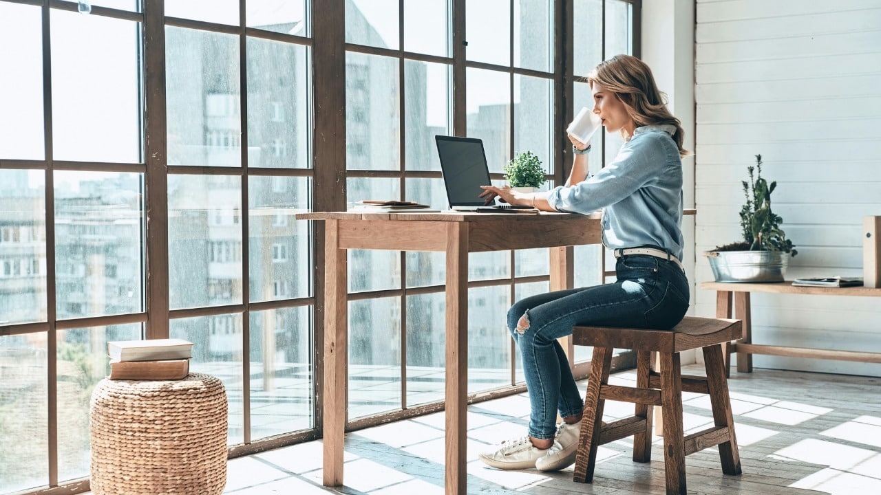A woman sits at a wooden desk by a large window, drinking from a mug.