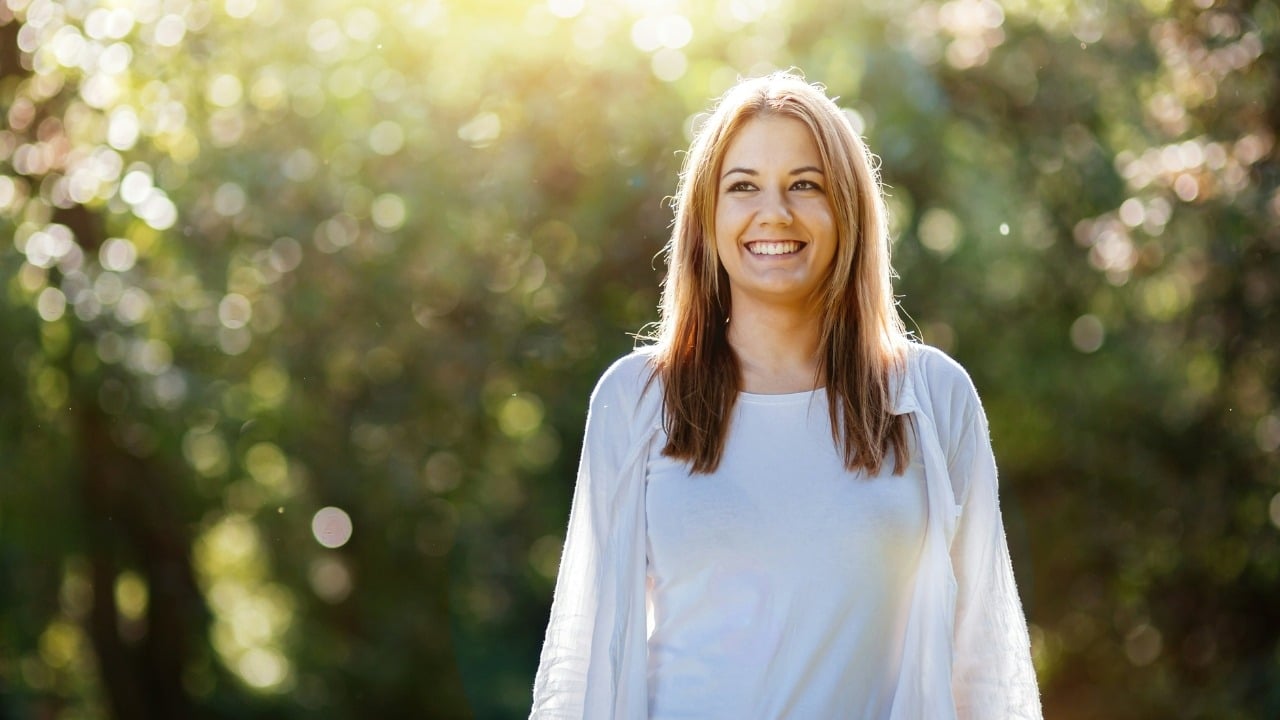 A smiling woman with blonde hair stands outdoors with sunlight filtering through trees behind her.
