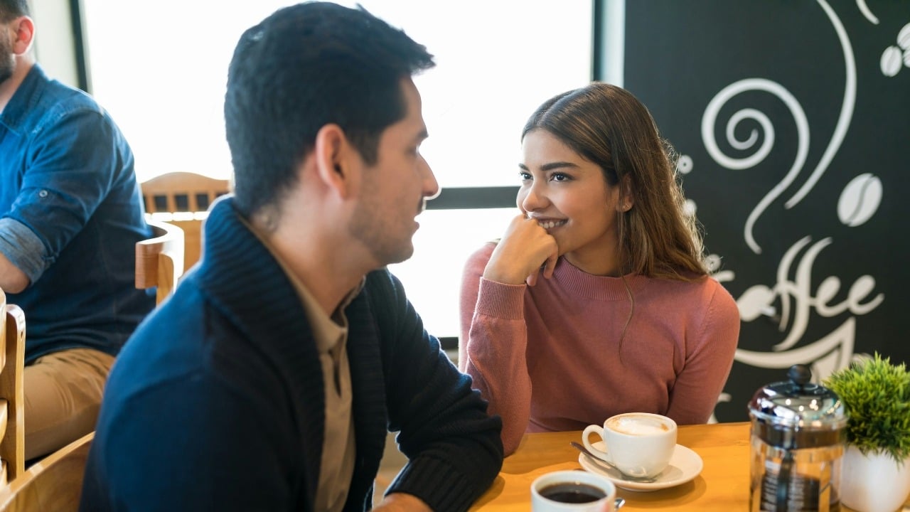 A man and woman sit at a cafe table with coffee cups, looking at each other.
