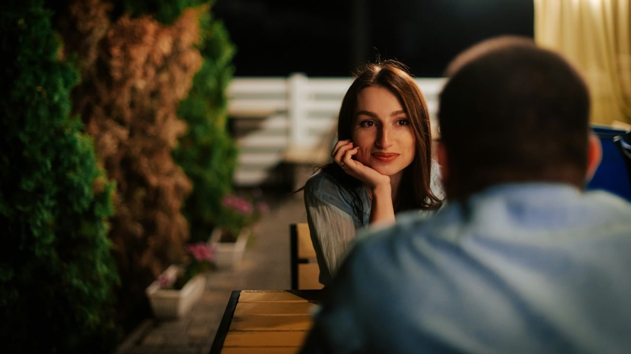 A woman rests her chin on her hand while looking at a man outdoors at night.