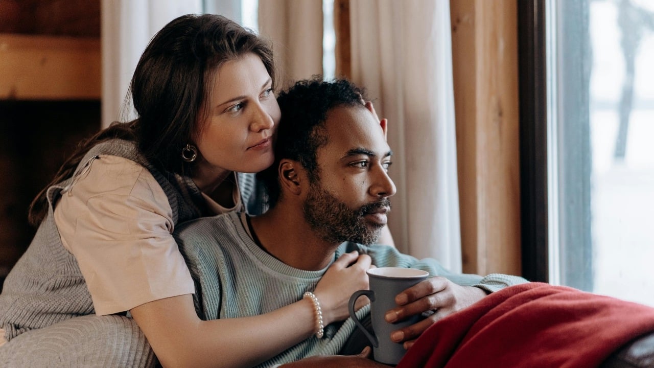 A woman leans against a man holding a mug as they look out a window.