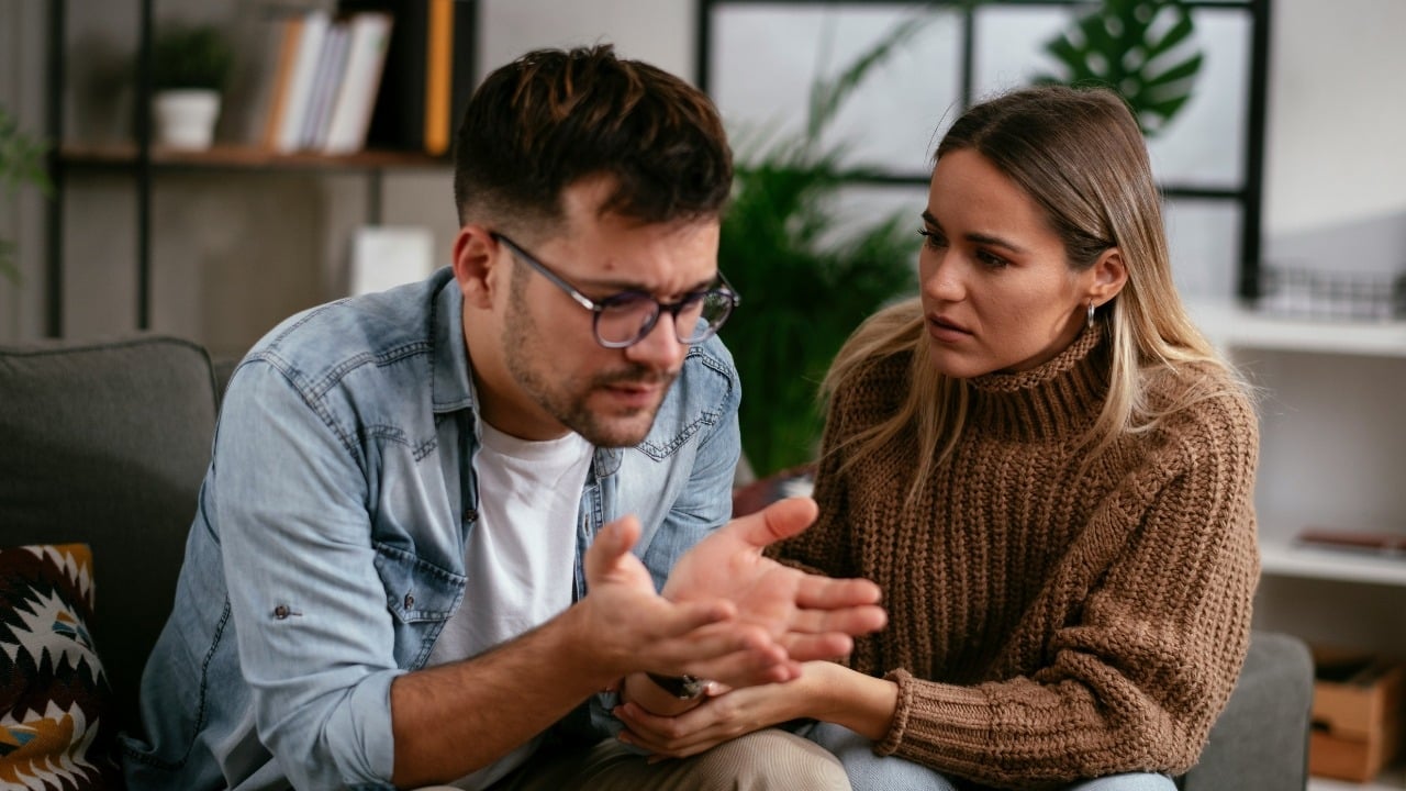 A man with glasses gestures with his hands while talking to a concerned-looking woman.