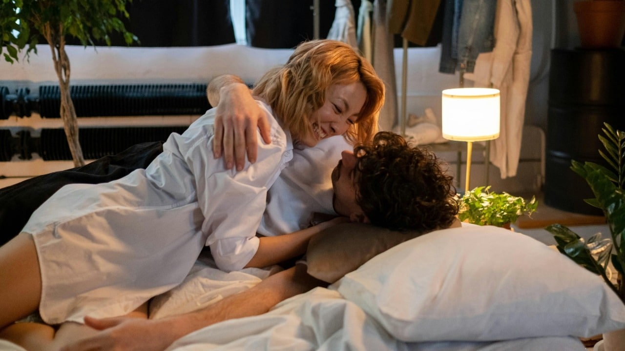 A woman in a white shirt smiles while leaning over a man on a bed.