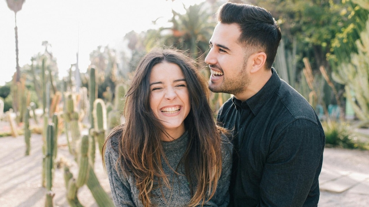 A man and woman laugh together outdoors with a background of various cacti plants.