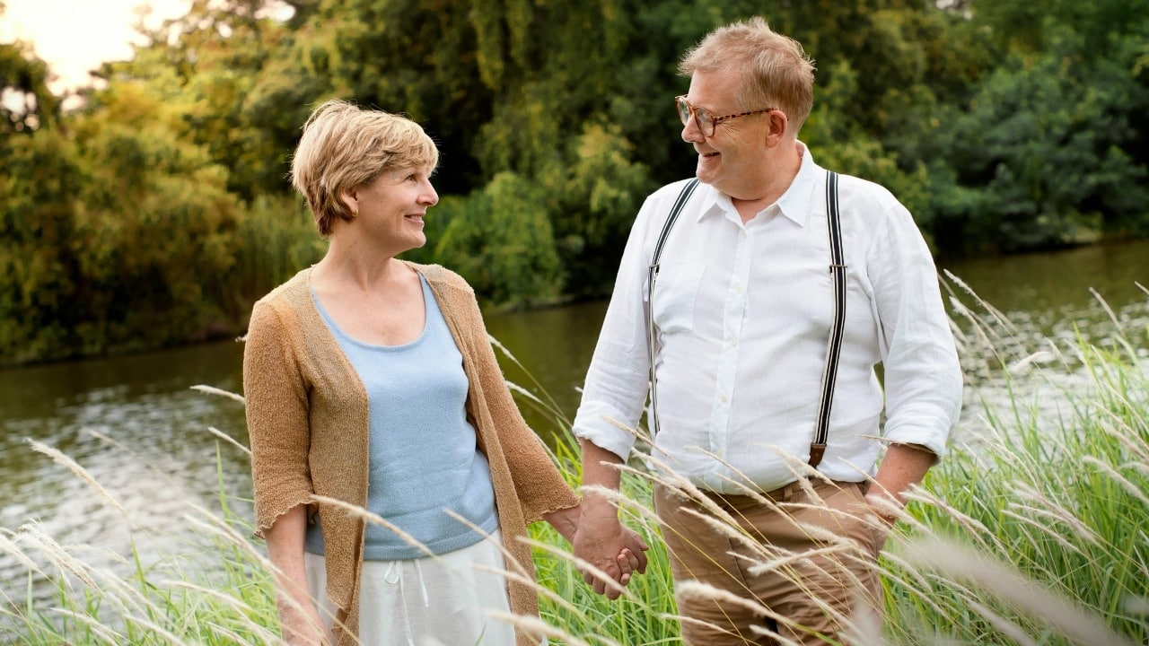 An older man and woman hold hands while standing in tall grass near a pond.