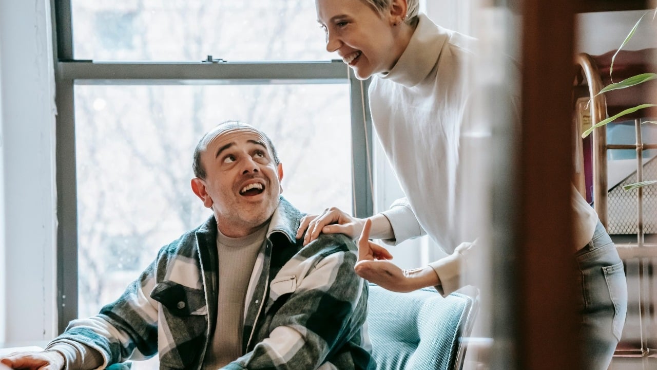 A woman stands and places a hand on the shoulder of a seated, smiling man.
