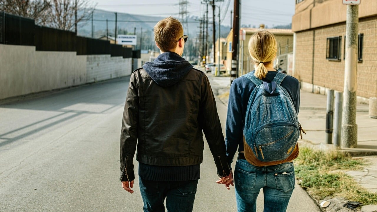 A man and woman seen from behind walk down a street while holding hands.