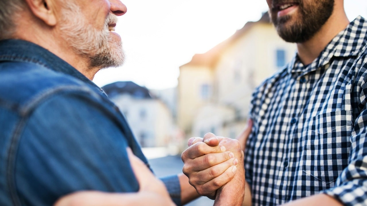 Two men in casual clothing are shown from the chest up, firmly shaking hands outdoors.