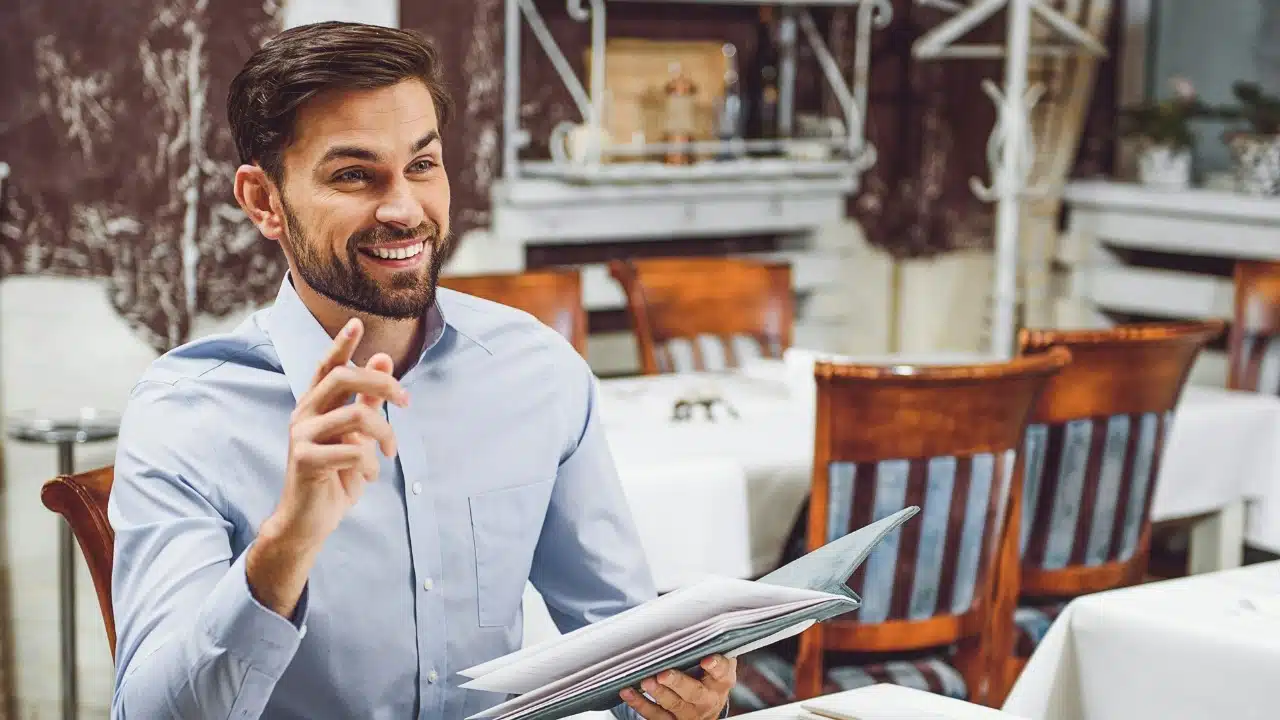 A smiling man in a light blue shirt holds a menu and points in a restaurant.