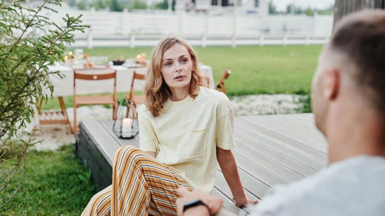 A woman in a yellow shirt and striped pants sits outdoors talking to a man.