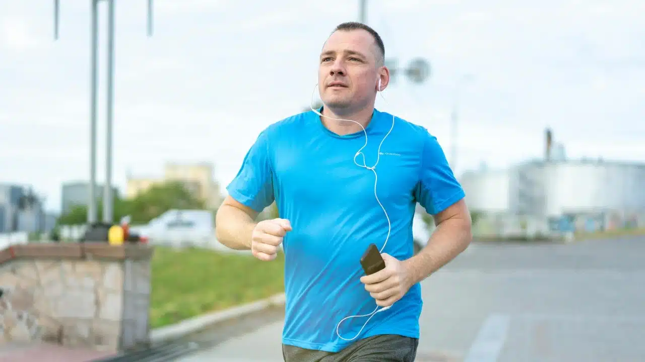 A man in a blue t-shirt runs outside while wearing earphones and holding a phone.