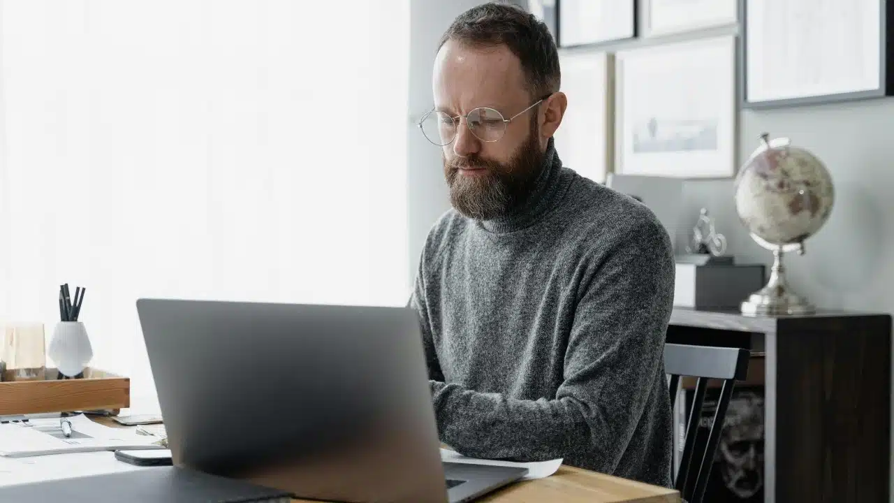 A bearded man with glasses and a grey turtleneck works on a laptop at a desk.