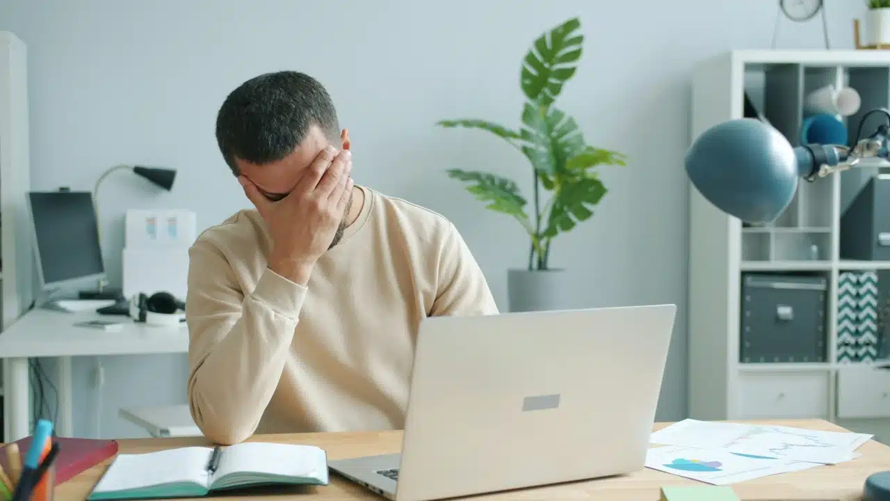 A man in a tan sweater sits at a desk with a laptop, covering his face.