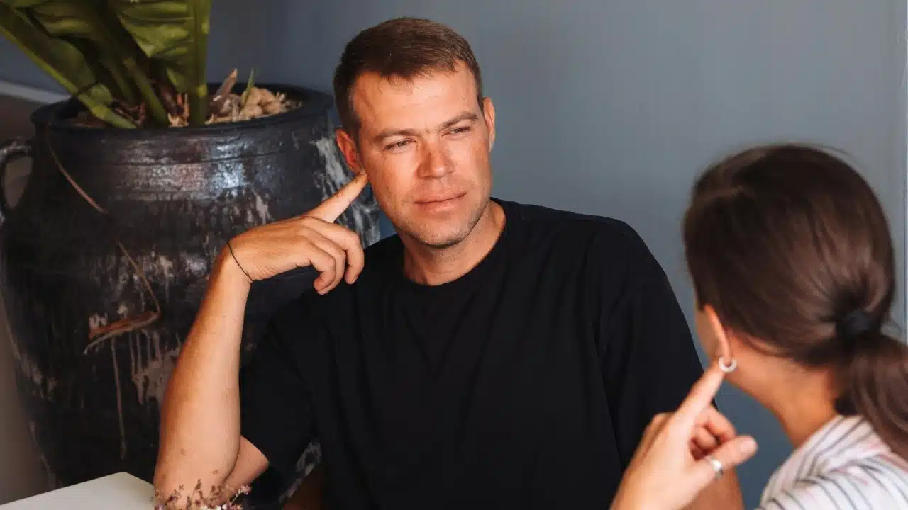 A man in a black shirt listens to a woman speaking in a seated indoor setting.