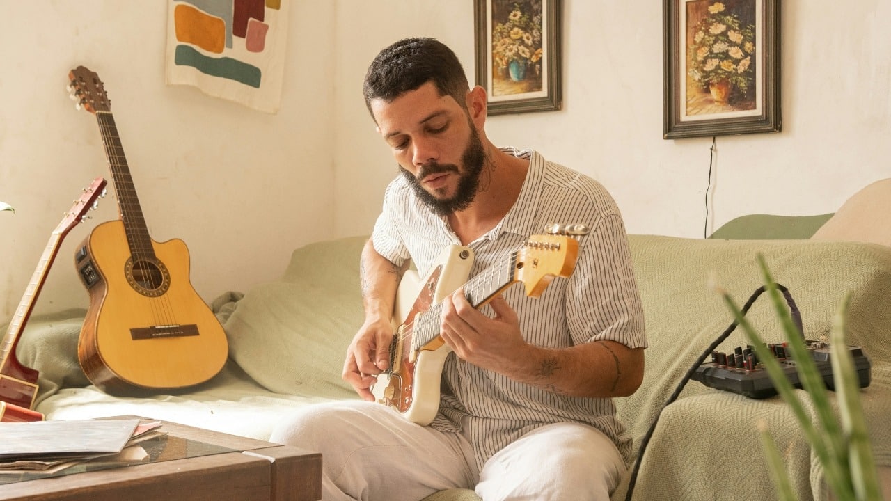 A man sits on a wooden floor playing an acoustic guitar in a bright room.