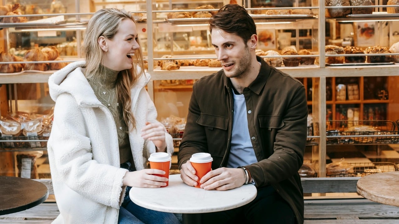 A man and woman sit at a round table outside a bakery holding coffee cups.
