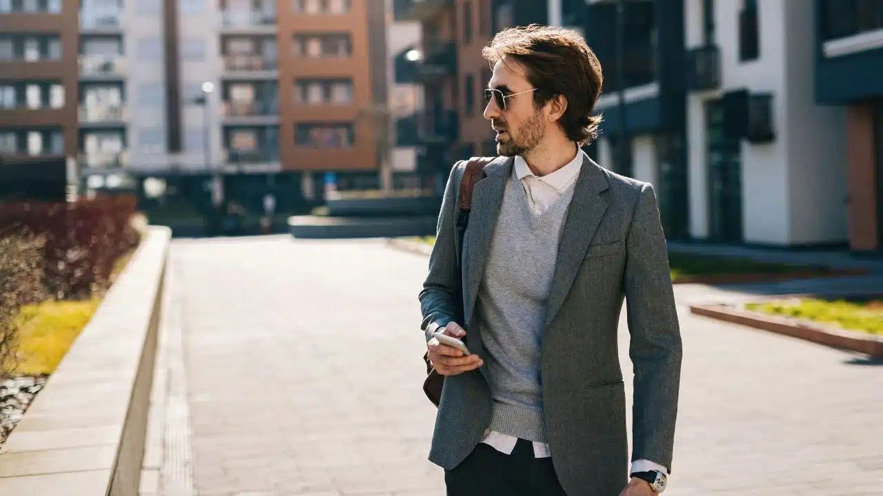 A man in a grey blazer and sunglasses walks outside while holding a mobile phone.