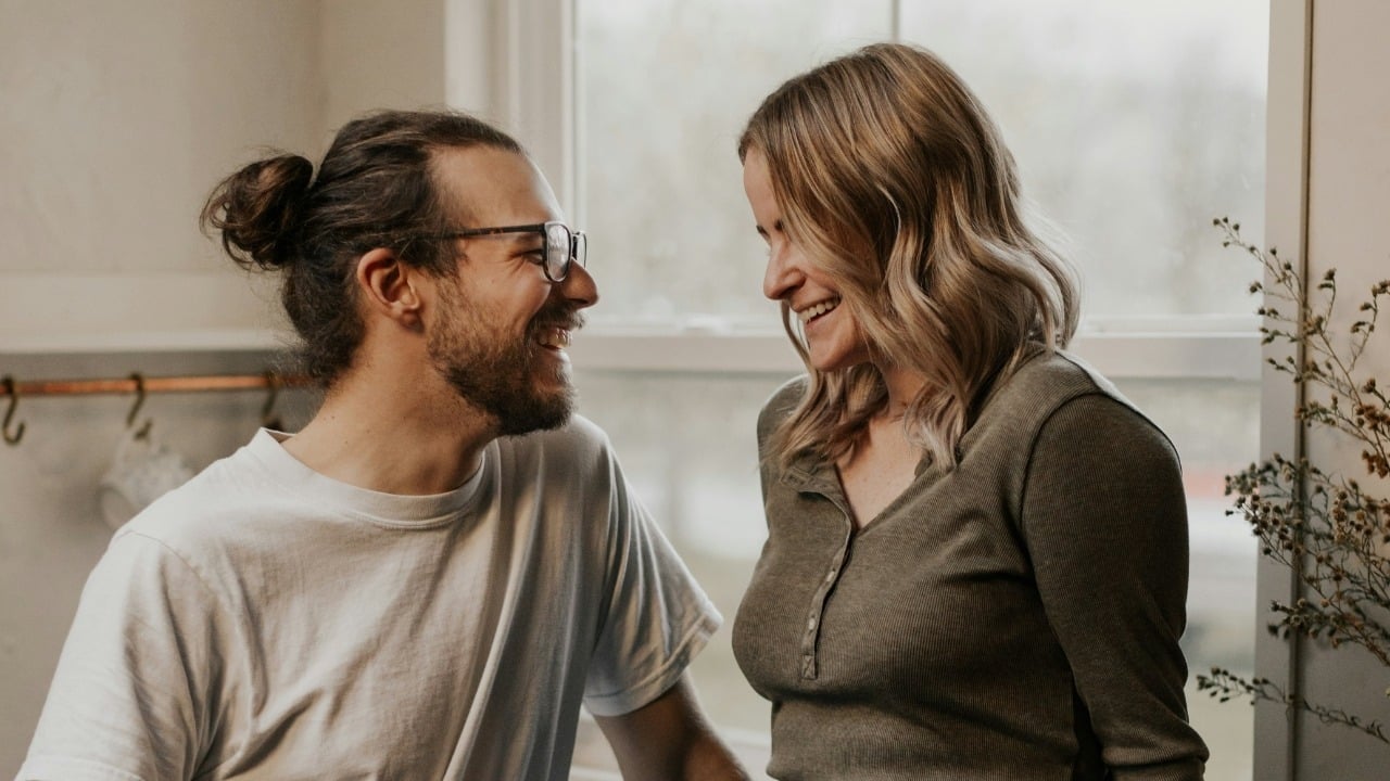 A man with a bun and a woman laugh while looking at each other indoors.