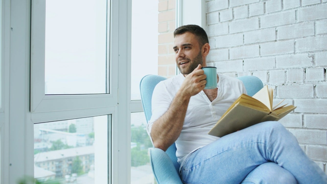 A man sits in a blue chair by a window, holding a mug and book.