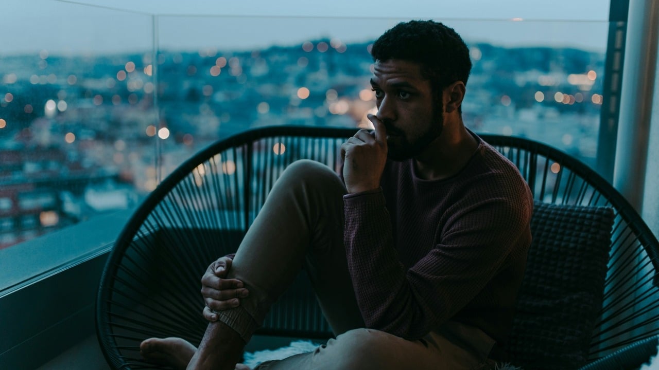 A man in a dark shirt sits on a sofa, looking calmly into the distance.