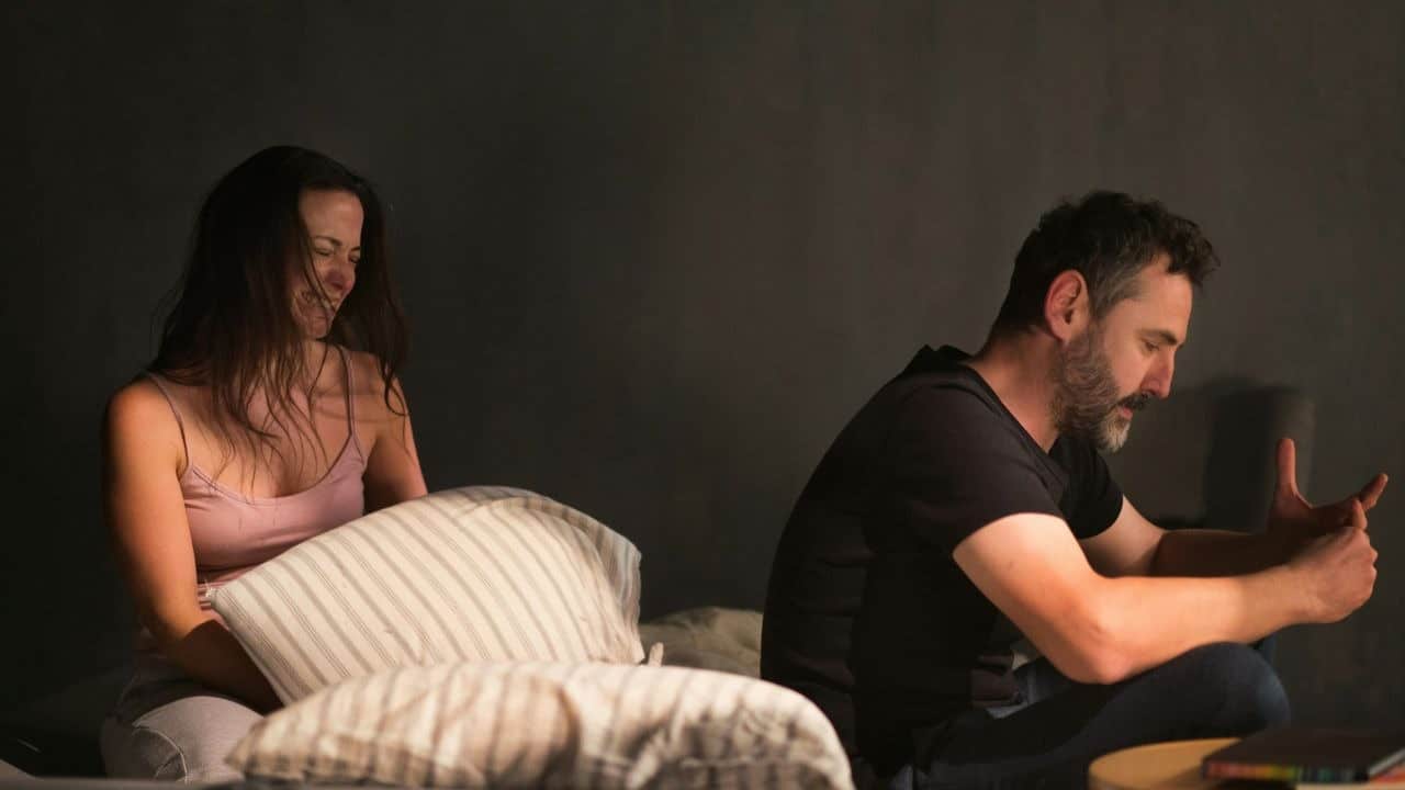 A woman and a man sit on a bed facing away from each other in a dark room.