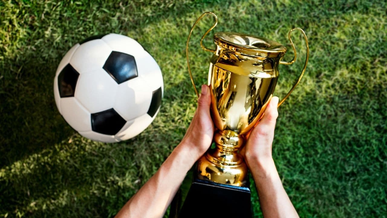 Hands hold a gold trophy cup on a grass field next to a soccer ball.
