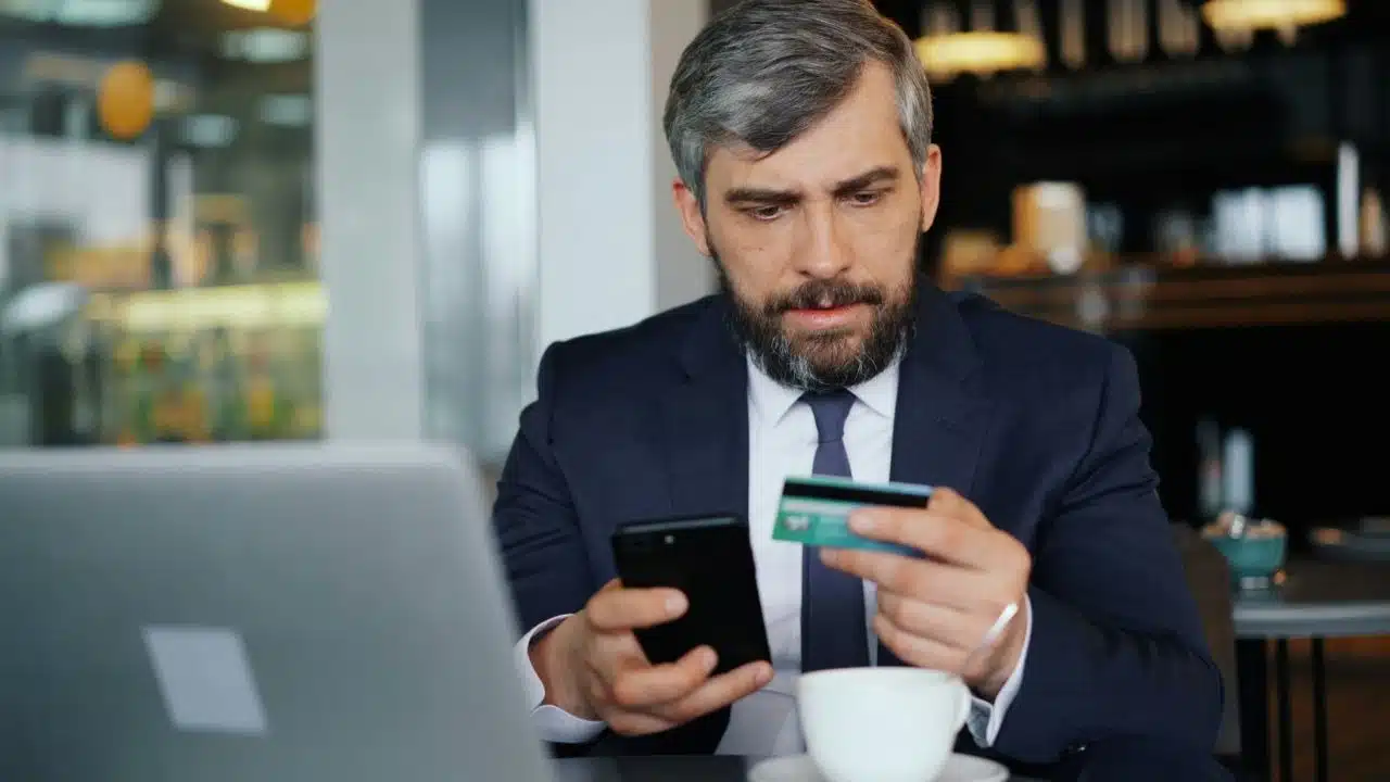 A bearded man in a suit looks at a credit card while holding a smartphone.