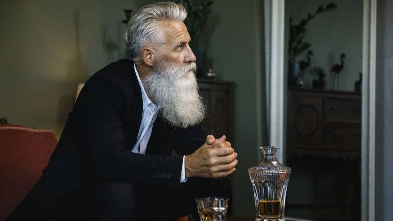 A man with a long white beard sits beside a glass decanter and a small glass.