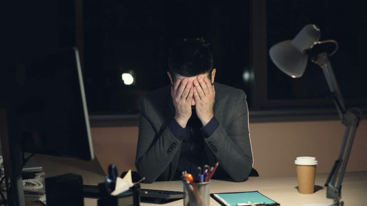 A man in a suit sits at a desk at night with his head in his hands.