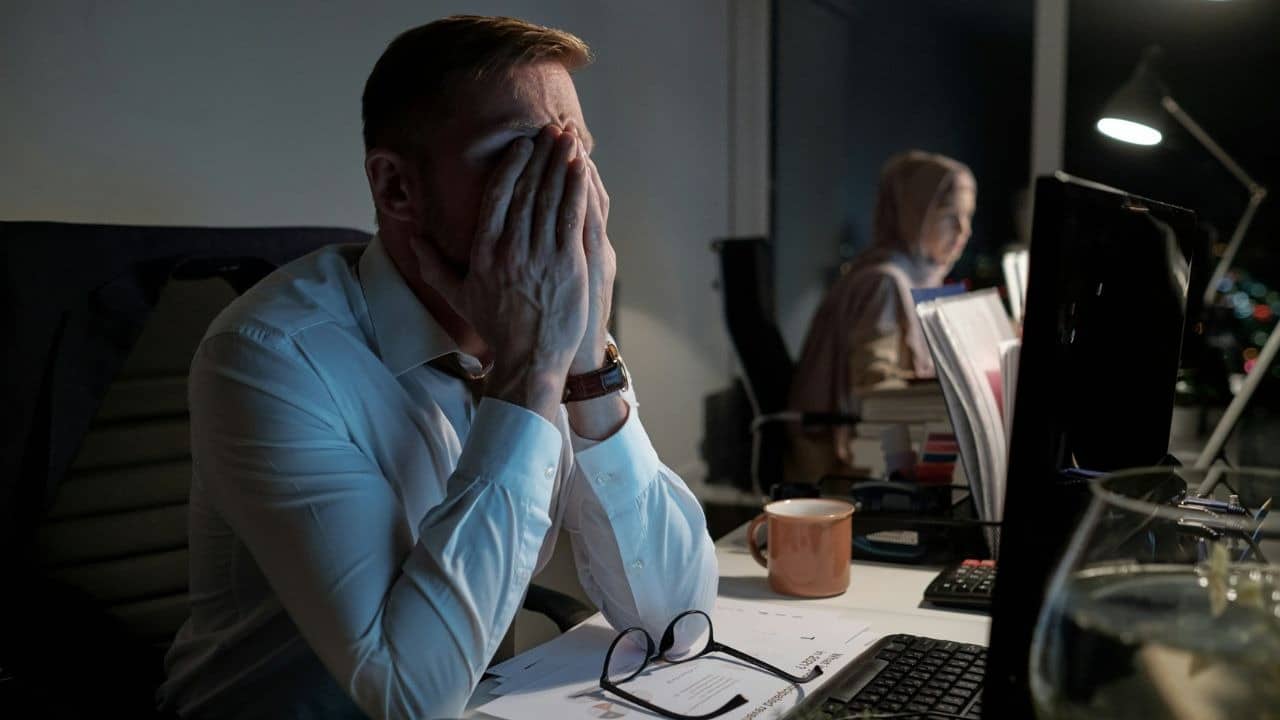 A man covers his face with his hands at a dark office desk at night.