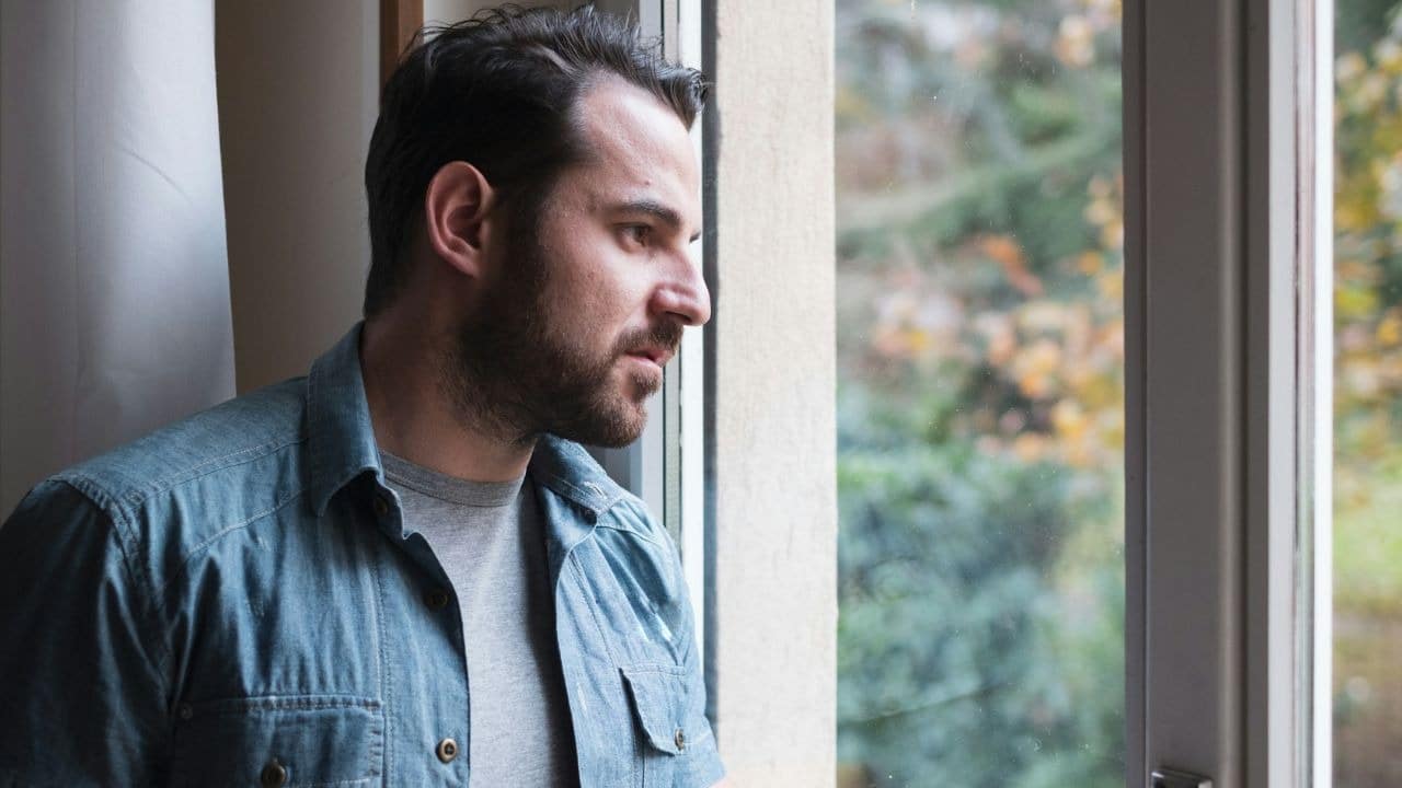 A bearded man in a denim shirt looks thoughtfully out of a window at trees.