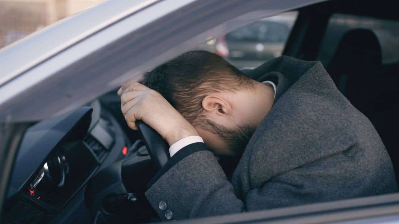 A man in a grey suit leans his head against the steering wheel of a car.