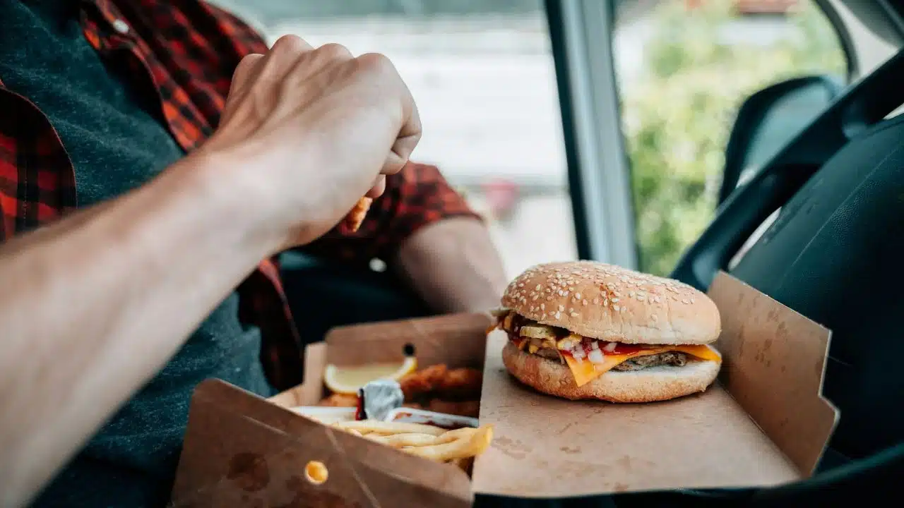 A person in a red plaid shirt eats a cheeseburger and fries inside a vehicle.