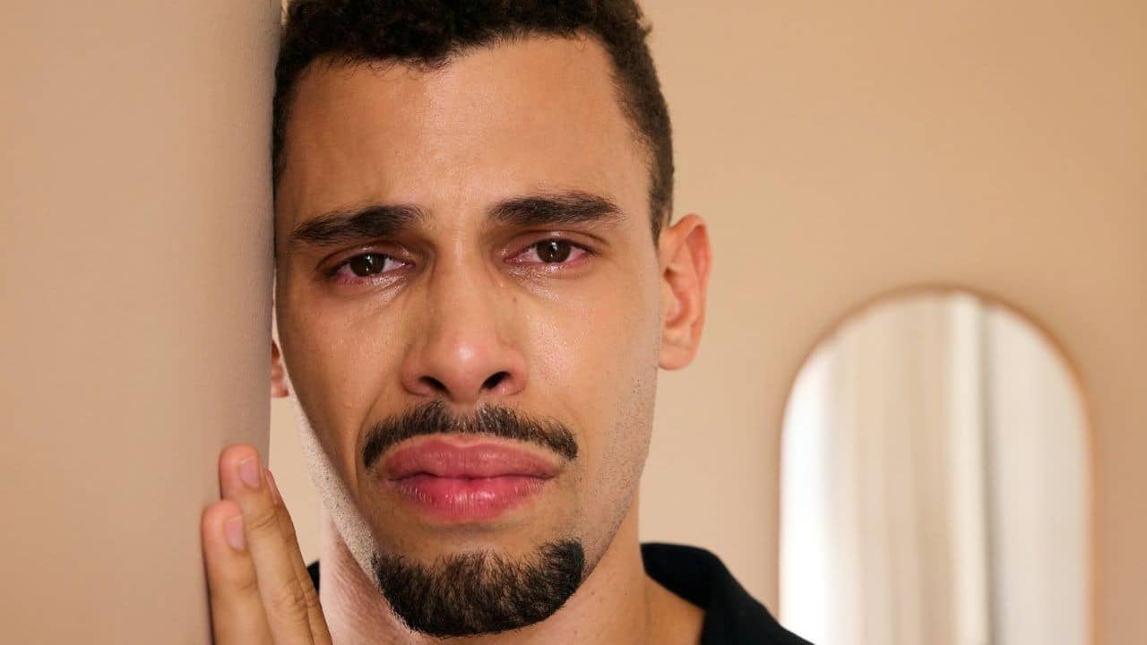 A man with a goatee leans his head against a wall with red, teary eyes.