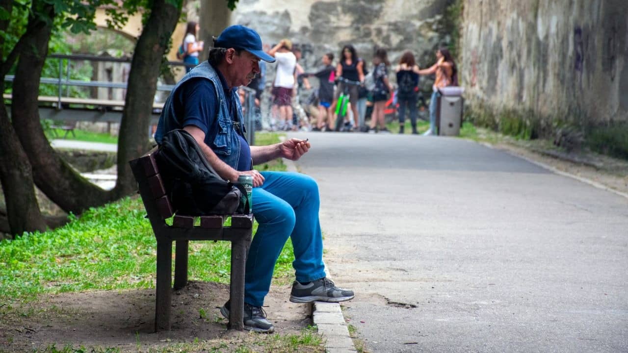 A man in a blue cap sits alone on a bench while others gather behind.