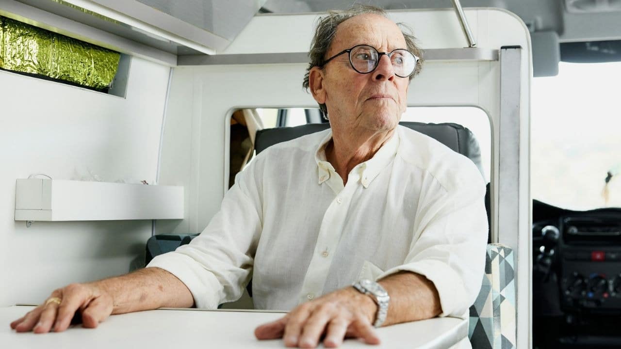 An older man wearing glasses and a white shirt looks out from inside a vehicle.