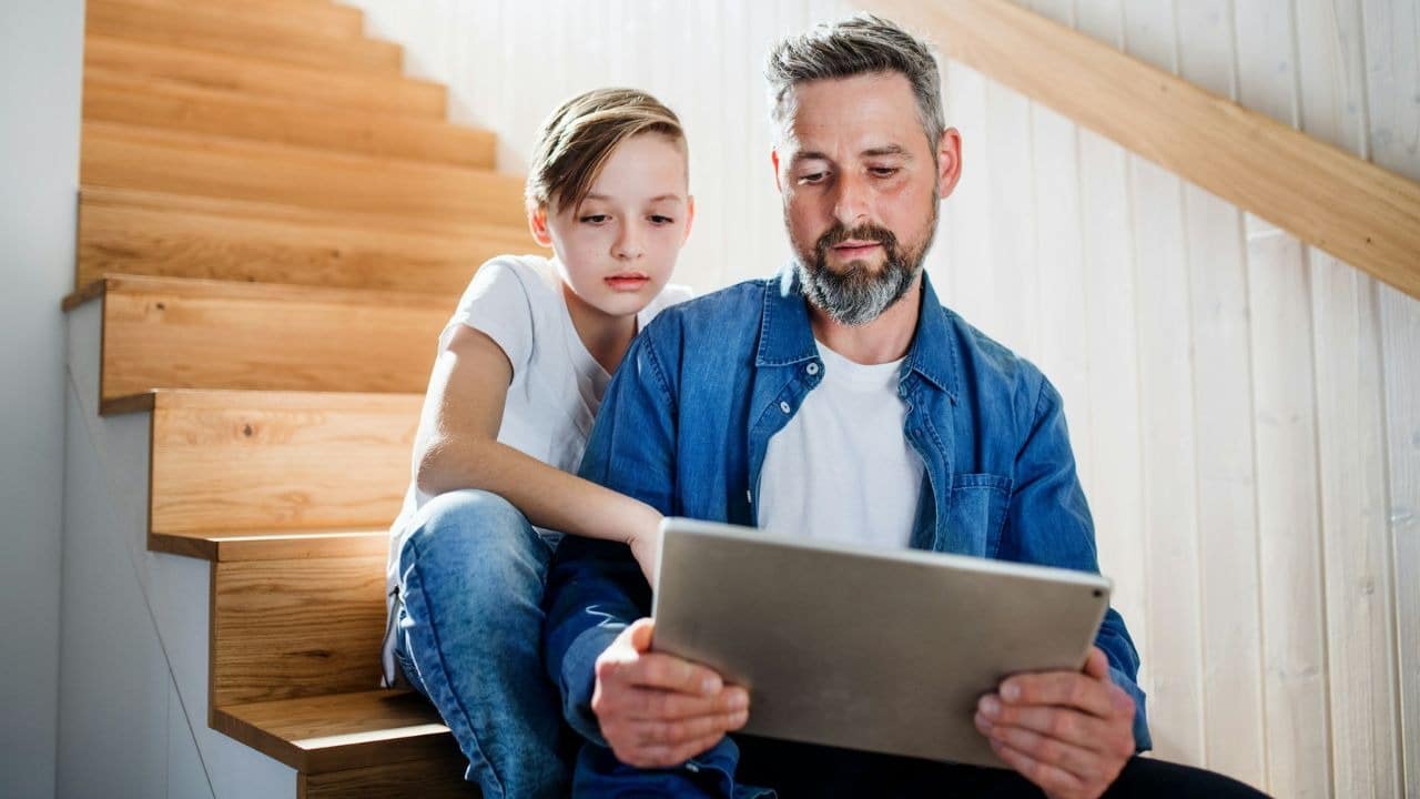 A man and a young boy sit on wooden stairs while looking at a tablet.
