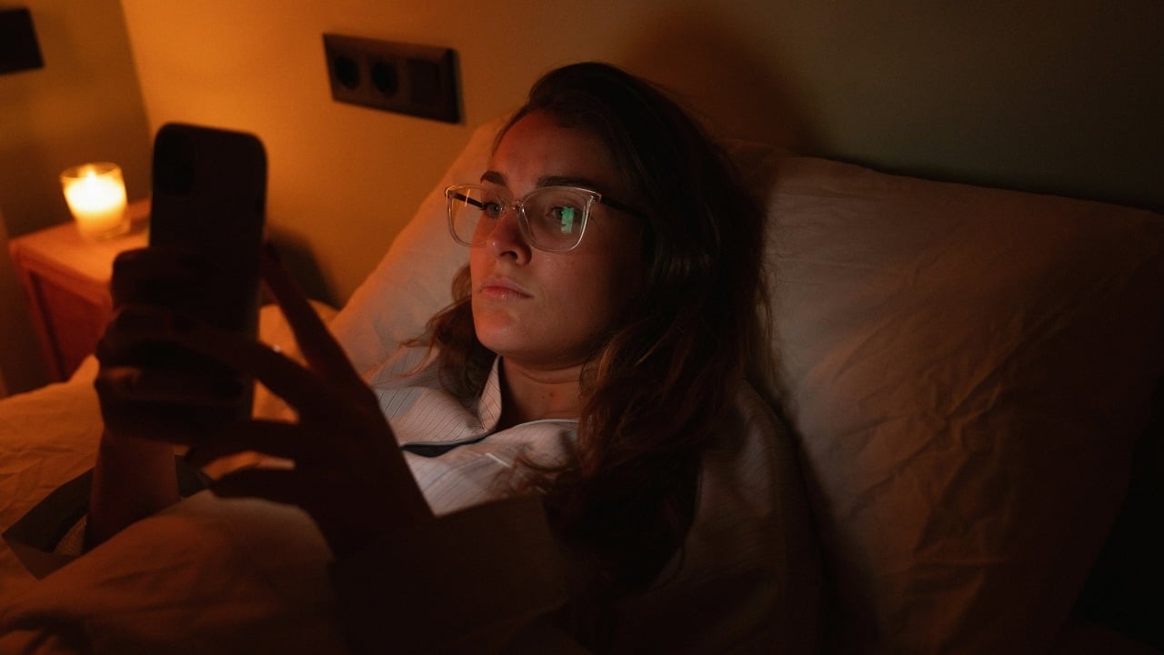 A person wearing glasses lies in bed at night looking at a glowing smartphone screen.